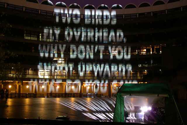 Projection on building reading: TWO BIRDS FLY OVERHEAD, WHY DON'T YOU SWOOP UPWARDS?